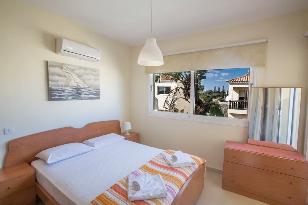 A cozy bedroom with wooden furniture, a sailboat painting, and a view of tropical greenery through the window, ideal for a relaxing vacation retreat.