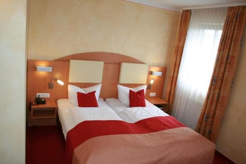 A cozy hotel room features a neatly made double bed with red accents, warm lighting from bedside lamps, and a window with patterned curtains, ideal for travelers seeking comfort and relaxation.