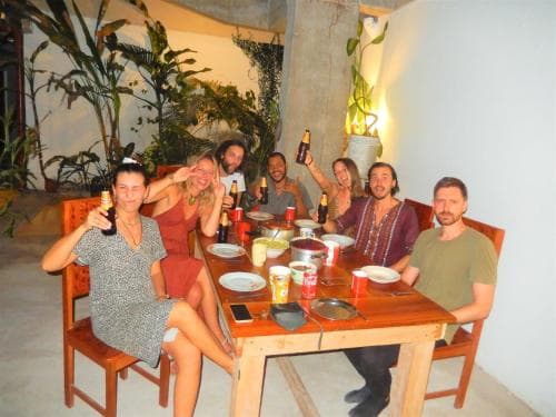 A lively group enjoys drinks and snacks around a rustic wooden table in a cozy indoor setting adorned with lush plants, perfect for a casual gathering or social event.