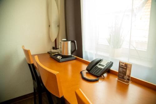 A cozy hotel room corner features a wooden desk with a kettle, telephone, and informational stand against a softly lit window, ideal for travelers seeking a quiet workspace or relaxation spot.