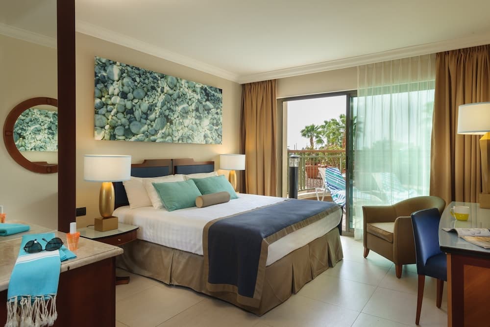 A cozy hotel room with a neatly made bed, vibrant artwork, and a balcony view of palm trees offers a serene retreat for travelers seeking relaxation.