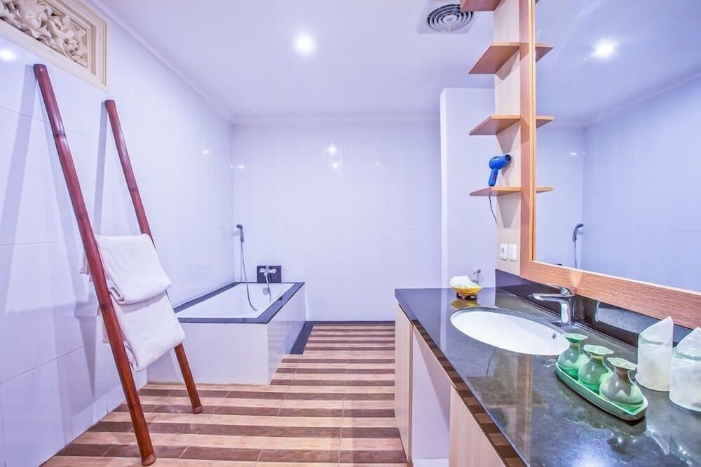 A serene hotel bathroom features a modern bathtub, sleek vanity with toiletries, wooden shelving holding a hairdryer, and a towel ladder against white tiled walls, ideal for travelers seeking comfort and style.