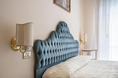 A cozy hotel room features a plush blue tufted headboard bed flanked by elegant wall sconces, creating a serene and inviting atmosphere ideal for travelers seeking comfort and relaxation.