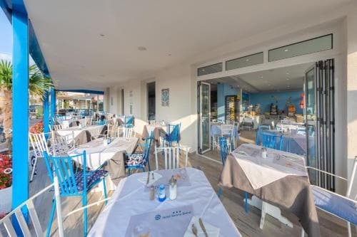 A serene coastal restaurant patio features neatly arranged tables with blue chairs under a bright sky, ideal for travelers seeking a relaxing dining experience by the sea.