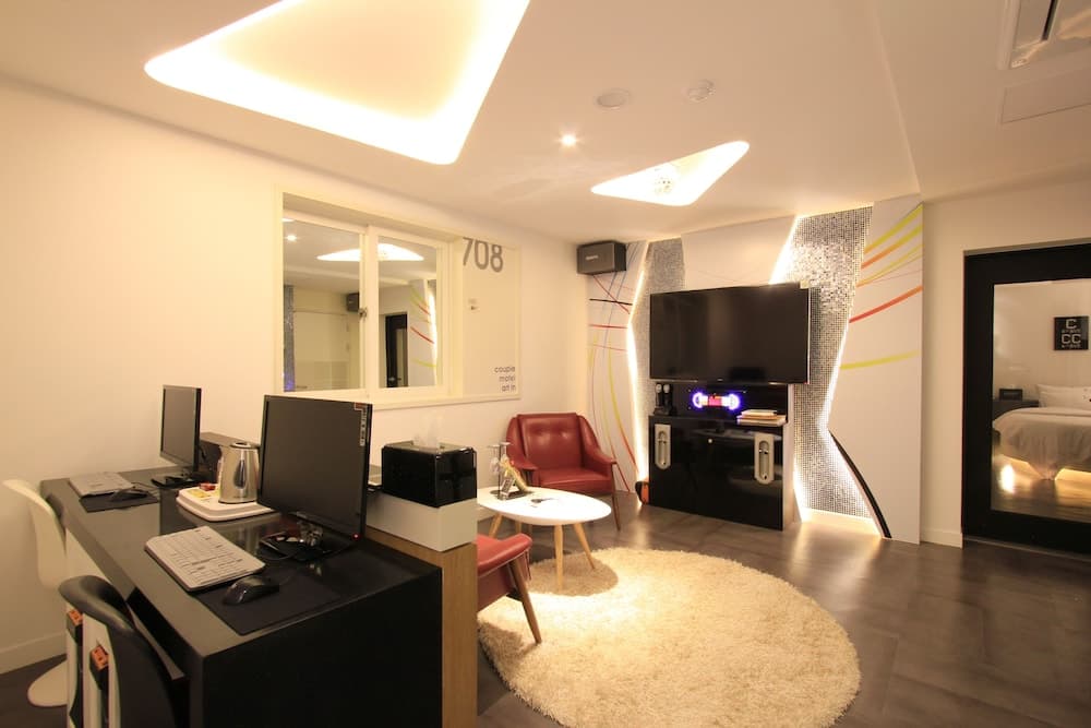 A modern hotel room features a cozy living area with a plush rug, sleek furniture, and a tech-equipped desk under soft ambient lighting, ideal for travelers seeking comfort and functionality.