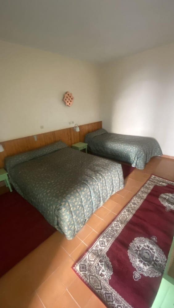 A cozy hotel room features two neatly made beds with patterned green bedding, a terracotta-tiled floor adorned with a decorative rug, and a simple wall-mounted light, ideal for travelers seeking a comfortable and budget-friendly stay.