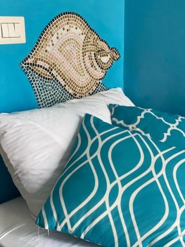 A cozy bedroom corner features a vibrant blue wall adorned with a mosaic conch shell headboard, complemented by patterned pillows, ideal for travelers seeking a serene and artistic retreat.