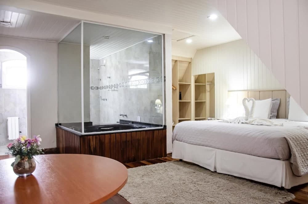 A cozy bedroom with a glass-enclosed bathroom, wooden bathtub, neatly made bed, and soft lighting creates an inviting atmosphere ideal for a relaxing getaway.