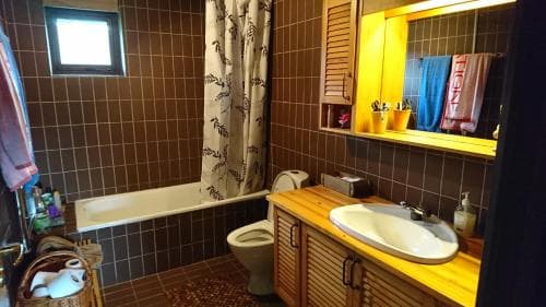 A cozy bathroom with brown tiled walls, wooden vanity, and a bathtub under a small window, exuding a warm and rustic charm ideal for travelers seeking a quaint accommodation experience.