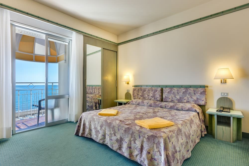 A cozy hotel room with a neatly made bed adorned in floral patterns, soft lighting, and a balcony offering a serene ocean view, ideal for travelers seeking relaxation by the sea.