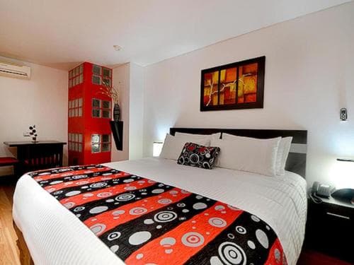 A cozy hotel room features a vibrant red and black patterned bedspread, modern artwork on white walls, and a stylish red partition, ideal for travelers seeking comfort and contemporary design.