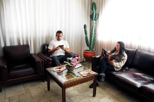A cozy living room scene features two individuals relaxing on leather sofas, surrounded by vibrant potted plants and a coffee table adorned with books and flowers, ideal for travelers seeking a tranquil retreat or a quiet reading spot.