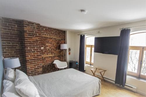 A cozy hotel room features an exposed brick wall, modern furnishings, and large windows letting in natural light, ideal for travelers seeking a blend of rustic charm and contemporary comfort.