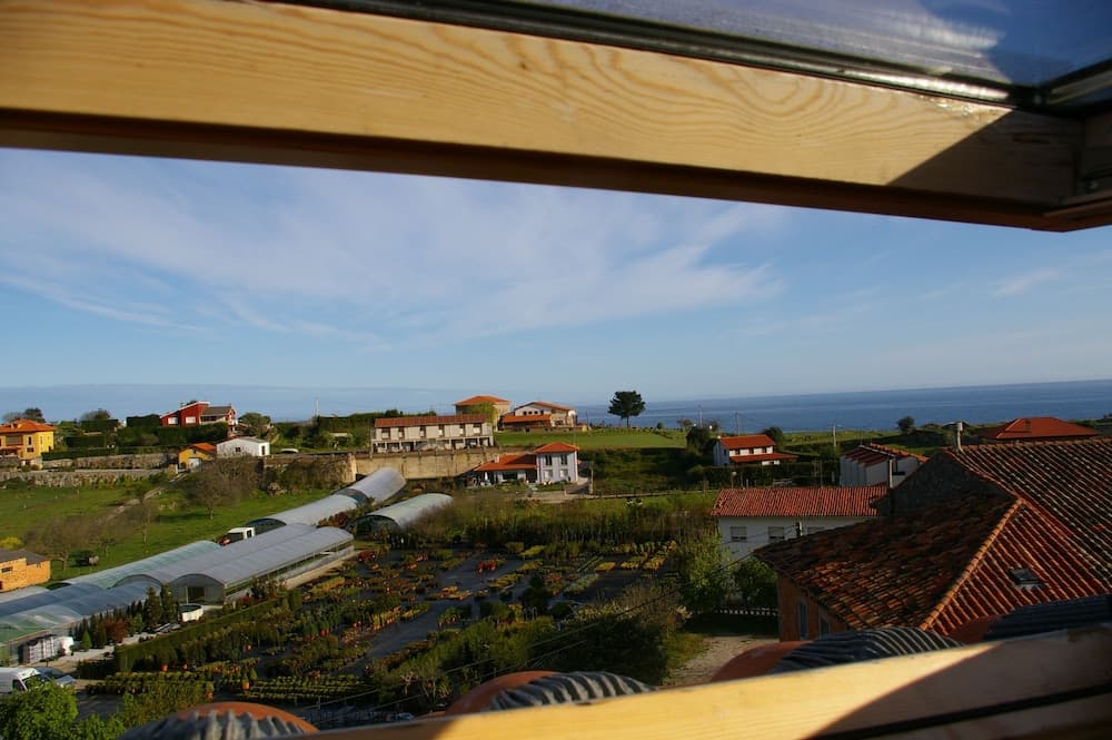 A serene coastal village view framed by wooden beams, featuring vibrant greenhouses, red-tiled roofs, and the calm ocean horizon, ideal for tranquil getaway inspiration.