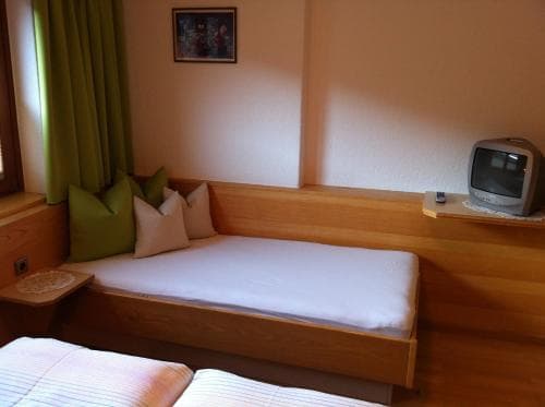 A cozy hotel room features a wooden daybed with pillows, a small TV on a shelf, and green curtains, creating a warm and inviting atmosphere ideal for travelers seeking comfort and relaxation.