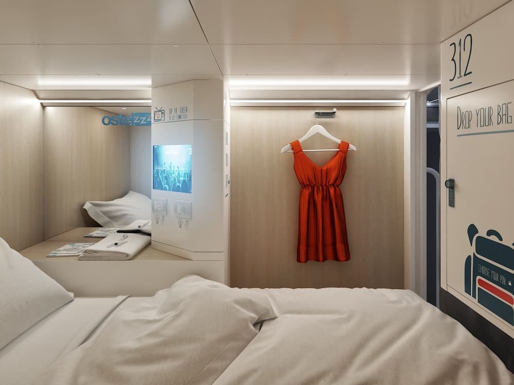 A cozy capsule hotel room features sleek wooden walls, a vibrant red dress hanging on the wall, and a built-in screen, ideal for travelers seeking modern comfort in compact spaces.