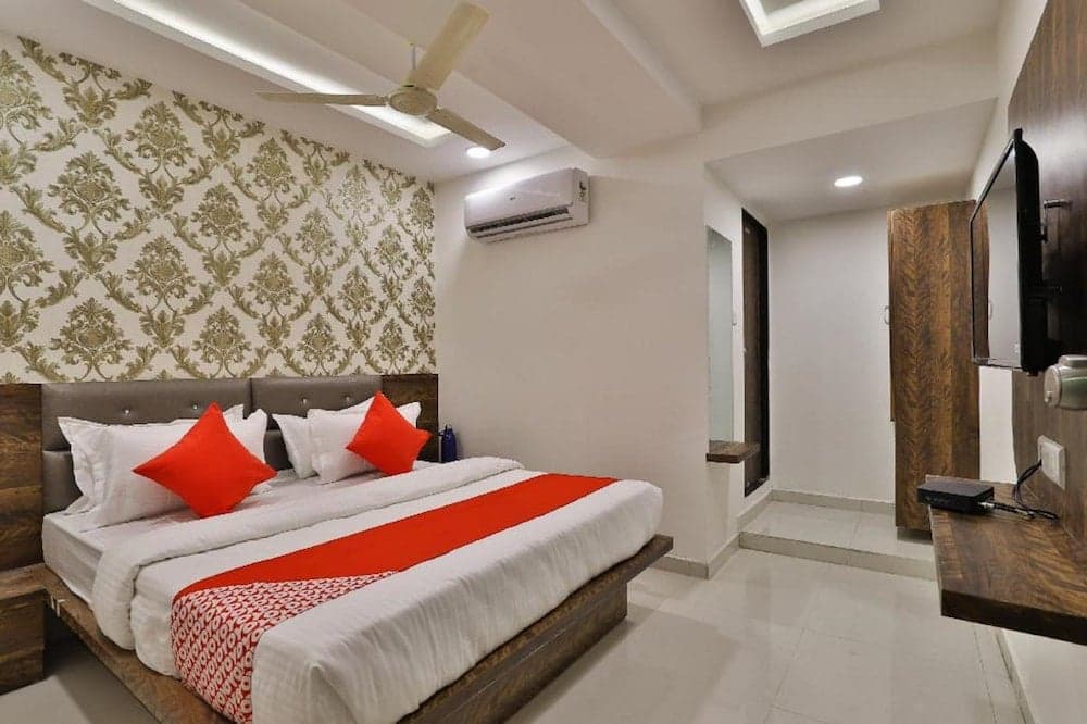 A cozy hotel room features a neatly made bed with red accents against a patterned wall, complemented by modern amenities like a mounted TV and air conditioning, ideal for travelers seeking comfort and convenience.