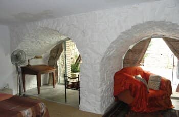 A cozy, rustic interior with white stone arches frames a serene living space featuring vibrant orange fabric, wooden furniture, and a fan, ideal for travelers seeking a tranquil retreat in a historic setting.