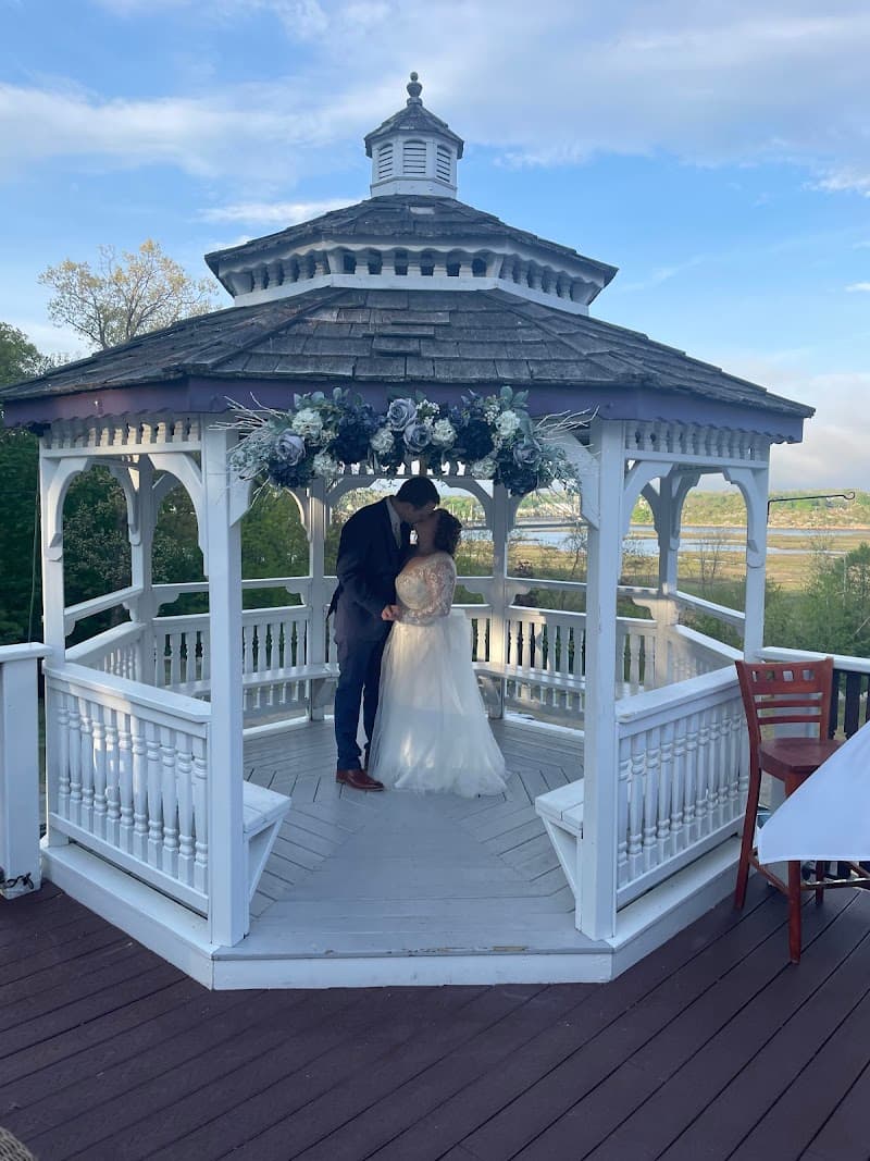A romantic couple shares an intimate moment under a beautifully adorned gazebo at the Castle Manor Inn in Gloucester, MA, set against a serene backdrop of trees and water, perfect for wedding photography and special occasions.