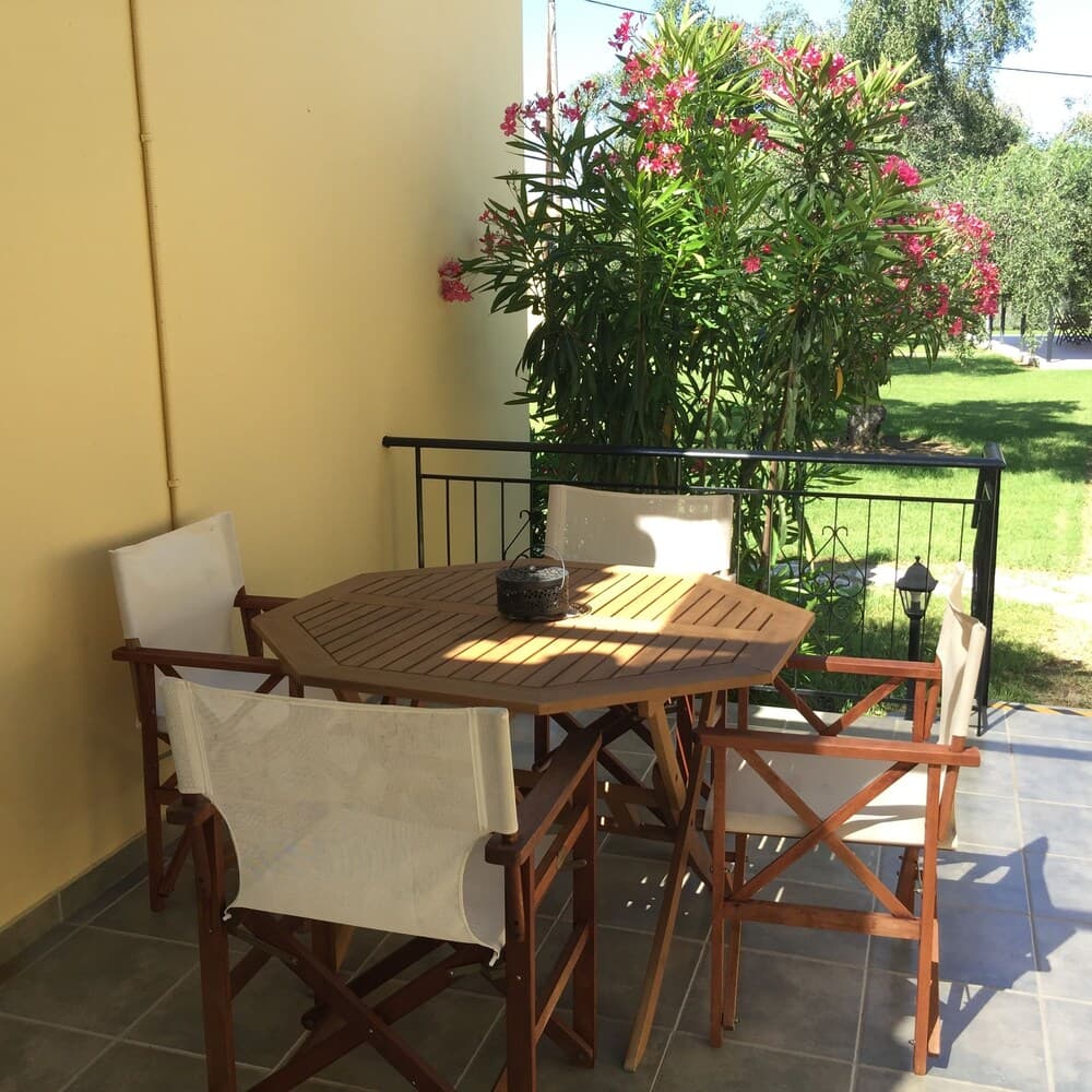 A cozy outdoor patio features a wooden table with chairs set against a yellow wall, surrounded by lush greenery and vibrant pink flowers, ideal for a relaxing vacation retreat.