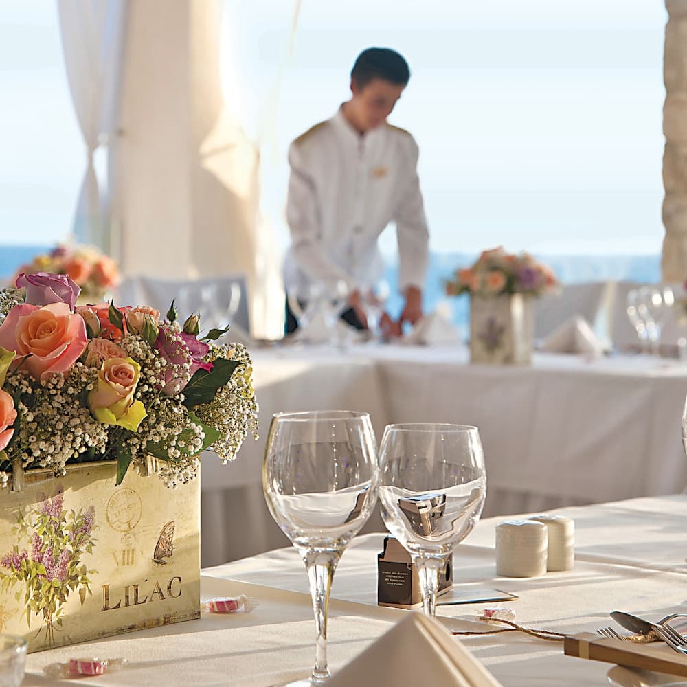 A serene seaside dining setup features elegant tables adorned with floral arrangements and wine glasses, creating an inviting atmosphere perfect for a luxurious beachside event or wedding reception.