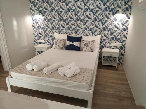 A cozy bedroom with a neatly made bed adorned with rolled towels, featuring vibrant blue leaf-patterned wallpaper, white bedside tables, and soft lighting, ideal for travelers seeking a comfortable and stylish accommodation.
