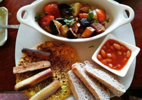 A hearty breakfast plate featuring scrambled eggs with sausages, toast slices, baked beans, and a colorful vegetable medley in a white dish, perfect for a cozy morning meal at a rustic café.