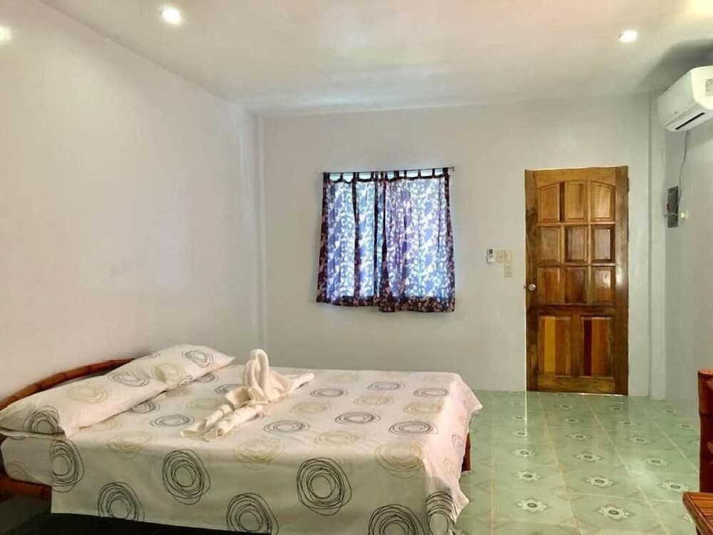 A cozy hotel room features a neatly made bed with patterned bedding, a wooden door, tiled flooring, and a window with floral curtains, ideal for budget travelers seeking comfort.