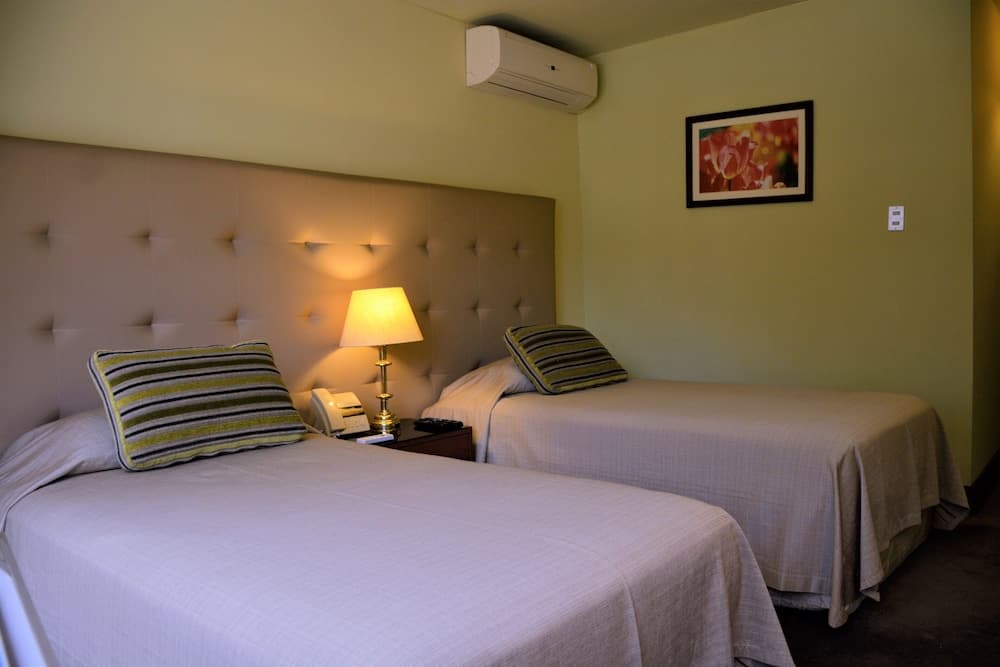 A cozy hotel room features two neatly made beds with striped pillows, a warm lamp on a nightstand, and a floral artwork, ideal for travelers seeking comfort and simplicity.