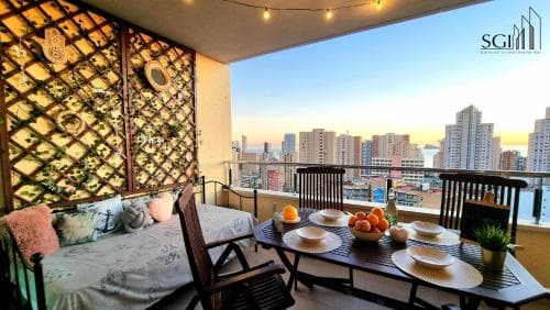 A cozy urban balcony adorned with string lights and greenery overlooks a vibrant cityscape at sunset, featuring a set dining table with fresh fruits, perfect for a relaxing getaway or city retreat experience.