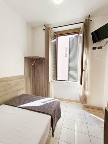 A cozy hotel room with tiled floors, a neatly made bed, wooden accents, and an open window letting in natural light, ideal for travelers seeking a simple and comfortable stay.