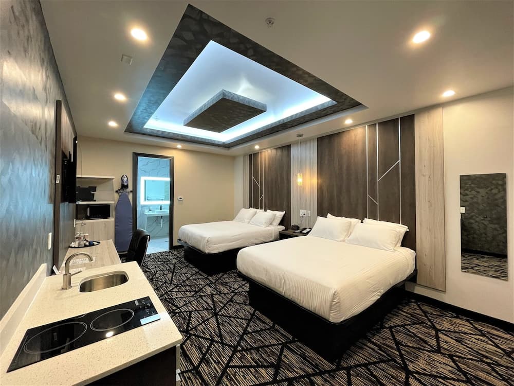 A modern hotel room features two cozy beds under a stylish ceiling light, a sleek kitchenette, and a bathroom visible through a glass door, ideal for travelers seeking comfort and convenience.
