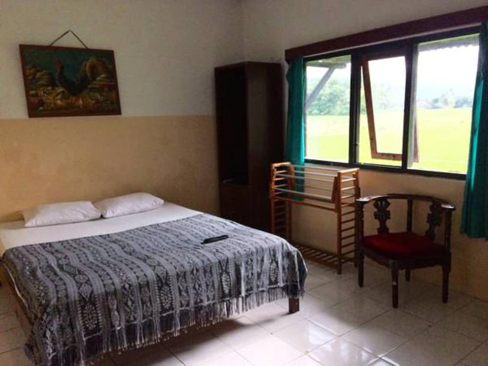 A cozy hotel room with tiled floors, a neatly made bed adorned with a patterned blanket, wooden furniture, and a window offering a serene view of greenery, ideal for travelers seeking a peaceful retreat.