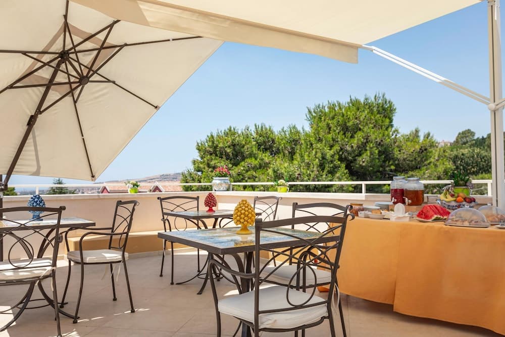 A serene rooftop terrace features cozy seating under large umbrellas, lush greenery in the background, and a vibrant breakfast spread, ideal for travelers seeking a relaxing morning retreat.