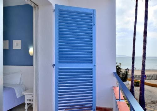A serene coastal hotel balcony features vibrant blue shutters overlooking a sandy beach with palm trees, ideal for travelers seeking relaxation by the sea.