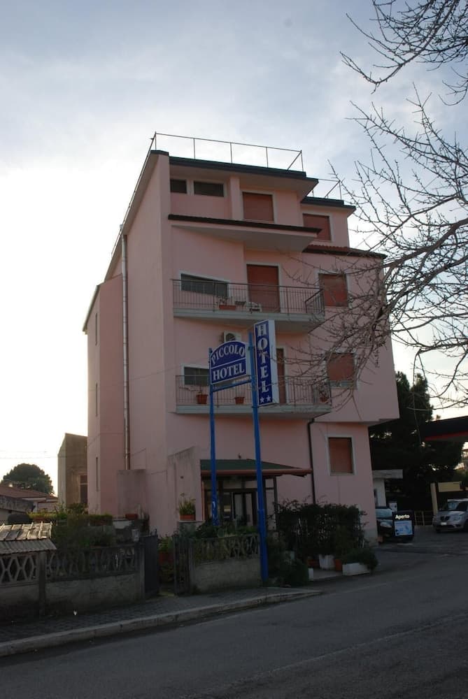 A quaint pink hotel with balconies and a "Piccolo Hotel" sign stands invitingly on a quiet street, ideal for travelers seeking cozy accommodations in a serene suburban setting.