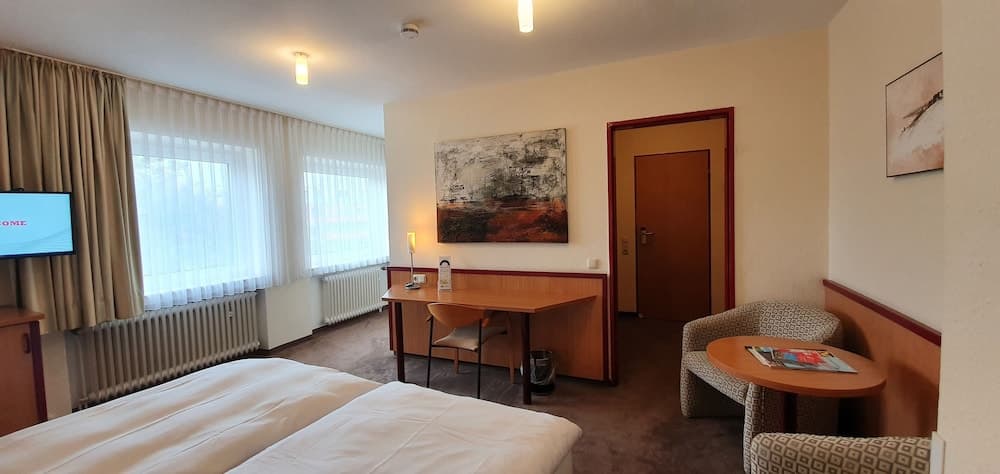 A cozy hotel room features twin beds, a wooden desk with a chair, a small seating area, and large windows with curtains, creating a comfortable and inviting atmosphere ideal for travelers seeking relaxation.