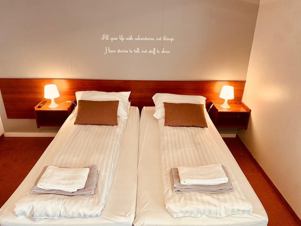 A cozy hotel room features two neatly made beds with folded towels, wooden bedside tables with lamps, and an inspirational quote on the wall, ideal for travelers seeking comfort and simplicity.