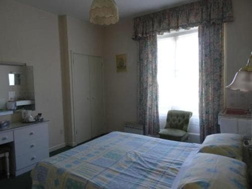 A cozy hotel room features a neatly made bed with a colorful quilt, floral curtains framing a bright window, and vintage furnishings, ideal for travelers seeking a comfortable and nostalgic stay.
