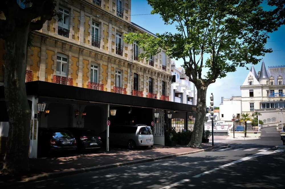 A serene European street scene features a charming yellow-patterned building with parked cars under its canopy, lush trees casting shade, and a distant castle-like structure, ideal for travelers seeking historic architecture and tranquil urban exploration.