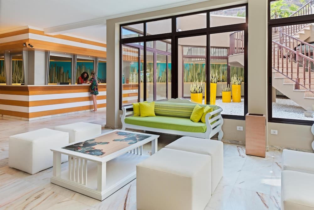 A vibrant hotel lobby features modern white furniture, green accents, and large windows overlooking lush greenery, creating an inviting atmosphere ideal for travelers seeking relaxation and comfort.