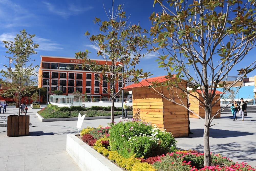 A vibrant urban plaza features colorful flower beds, wooden kiosks, and a modern hotel under a clear blue sky, ideal for city exploration and leisurely strolls.