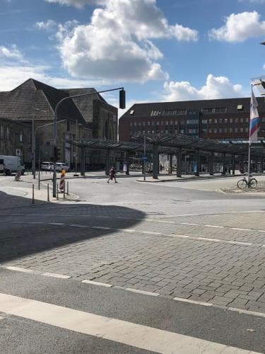 A spacious urban square under a partly cloudy sky features modern architecture, a covered pedestrian area, and a cyclist, ideal for city exploration and leisurely strolls.