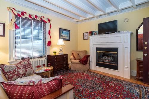 A cozy living room with warm yellow walls, a fireplace, a patterned rug, and a window with red curtains creates an inviting atmosphere perfect for a relaxing getaway.