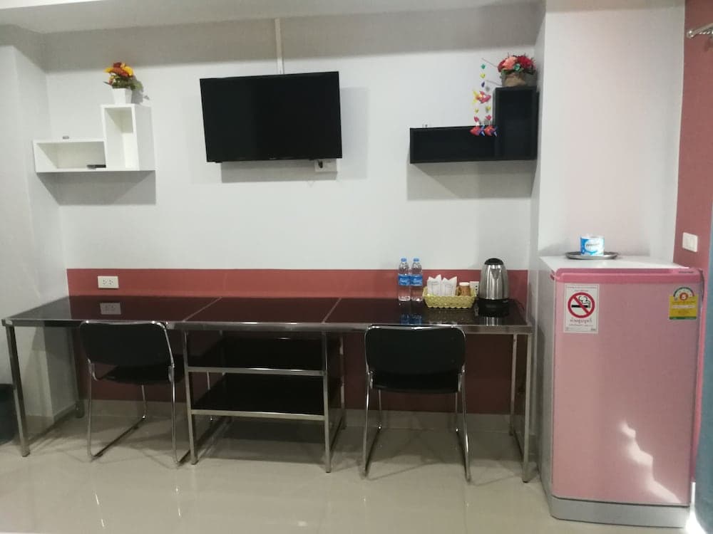 A cozy hotel room corner features a sleek dining table with chairs, a mounted TV, decorative shelves with flowers, a pink mini-fridge, and a kettle setup, ideal for travelers seeking comfort and convenience.