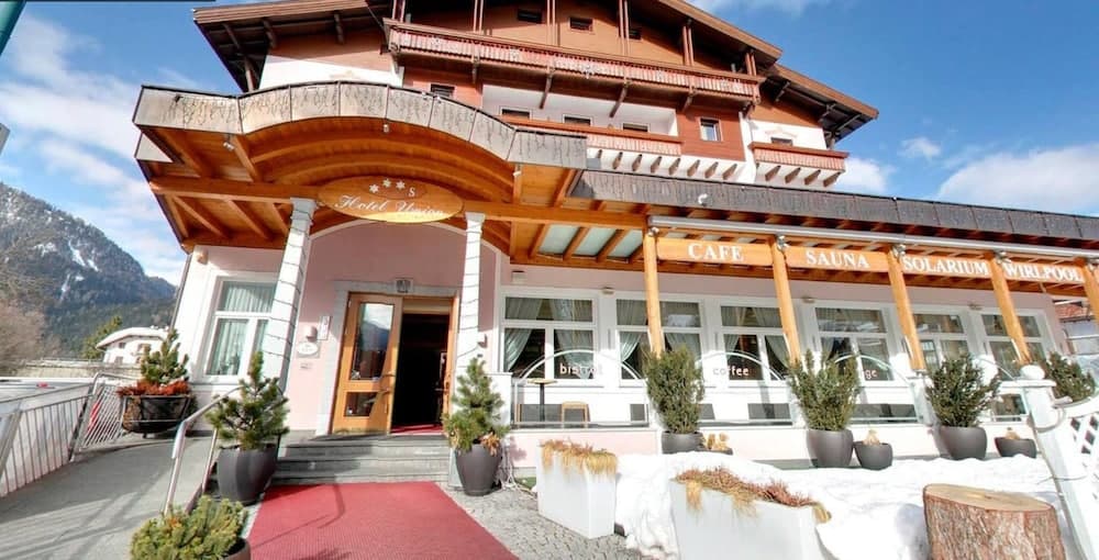 A cozy alpine hotel entrance with a red carpet, wooden beams, and snow-covered surroundings invites travelers to enjoy its café, sauna, solarium, and whirlpool amenities in a serene mountain setting.