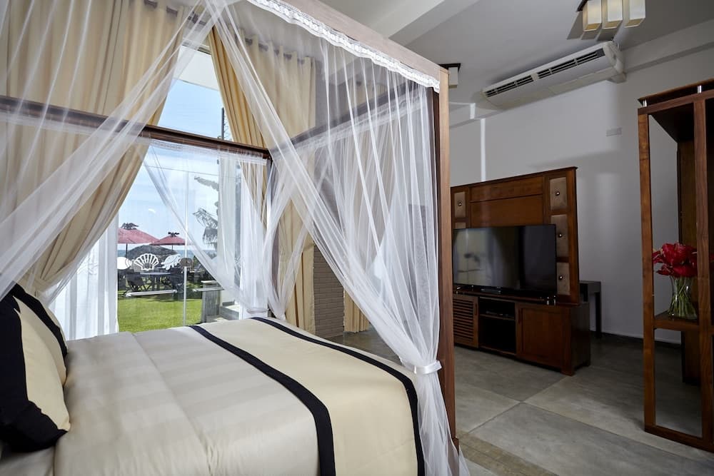 A serene hotel room features a cozy bed with mosquito netting, a view of lush greenery through large windows, and modern amenities like a flat-screen TV, ideal for travelers seeking comfort and relaxation.