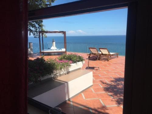 A serene coastal terrace framed by a doorway offers a breathtaking ocean view, complete with lounge chairs, potted plants, and a tranquil ambiance ideal for a relaxing getaway.