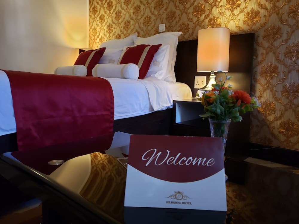 A cozy hotel room with elegant gold-patterned wallpaper, a neatly arranged bed with red accents, and a welcoming sign on the desk creates an inviting atmosphere perfect for travelers seeking comfort and luxury.