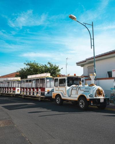 A charming open-sided tourist train navigates a sunny street beside white buildings under a clear blue sky, ideal for leisurely sightseeing tours.